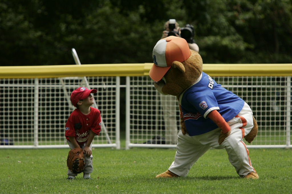 A tee ball player smiles at the mascot in the out field.