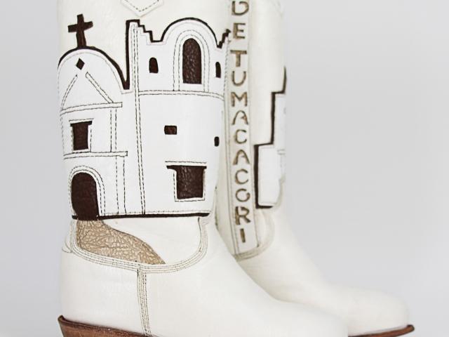 Miniature white leather boots with cutout name, "San Cayentano de Tumacacori," leatherwork depiction of the mission, and a gold leather cord for hanging. Signed inside by master craftsman Paul Bond, AZ, of the Paul Bond Boot Company.