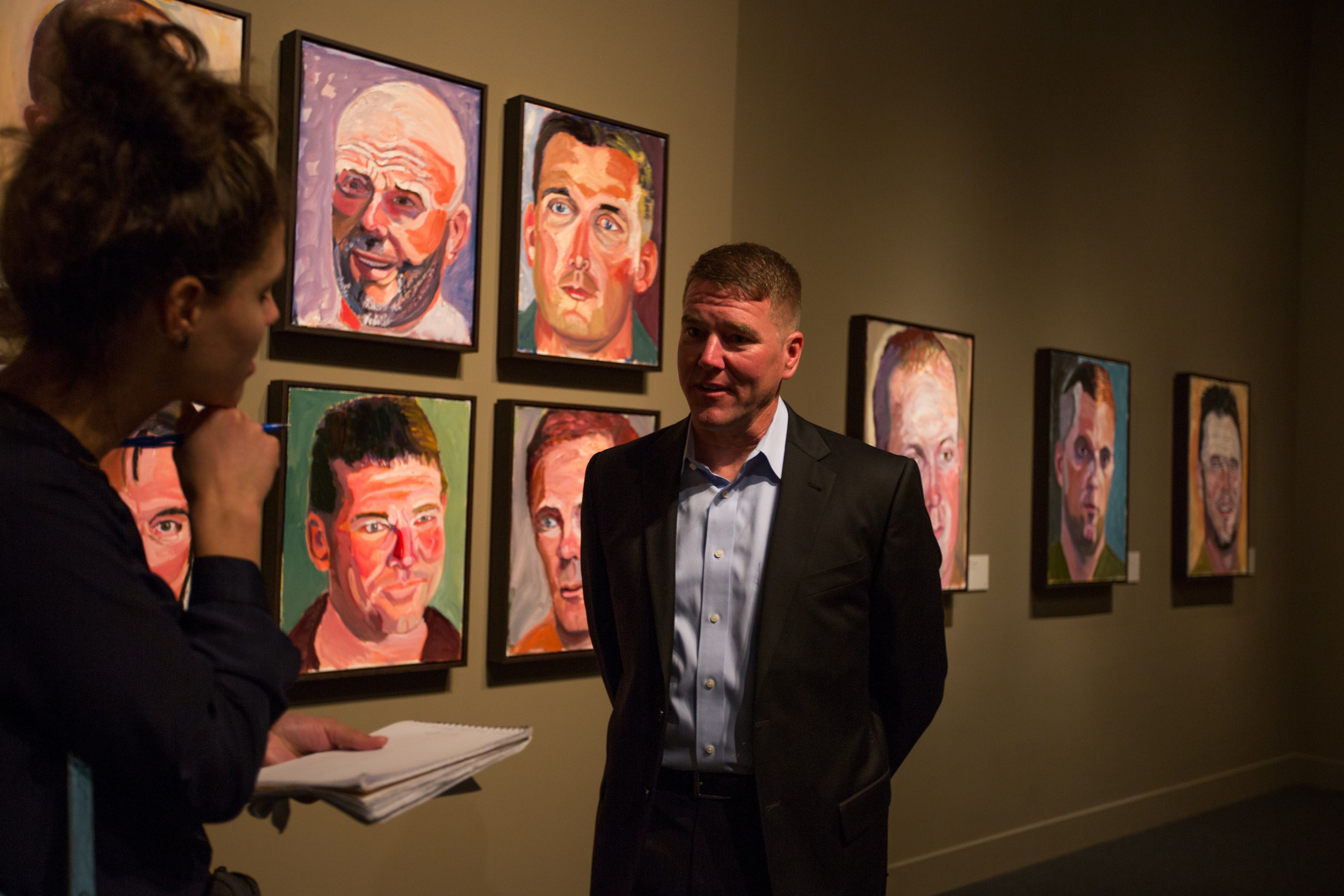 A photo of a man being interviewed by a woman as he stands near his portrait painted by President George W Bush, part of the 2017 exhibit Portraits of Courage, at the George W Bush Library and Museum
