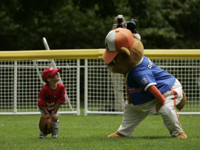 A tee ball player smiles at the mascot in the out field.