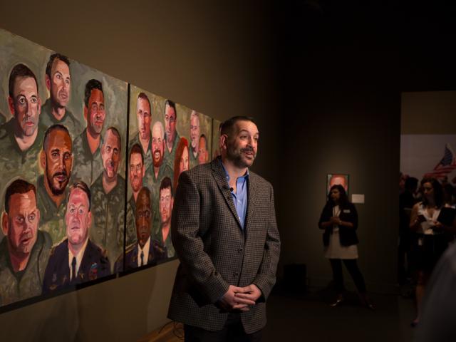 A photo of a man being filmed near his portrait and others painted by President George W Bush, part of the 2017 exhibit Portraits of Courage, at the George W Bush Library and Museum
