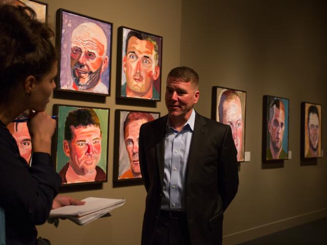 A photo of a man being interviewed by a woman as he stands near his portrait painted by President George W Bush, part of the 2017 exhibit Portraits of Courage, at the George W Bush Library and Museum