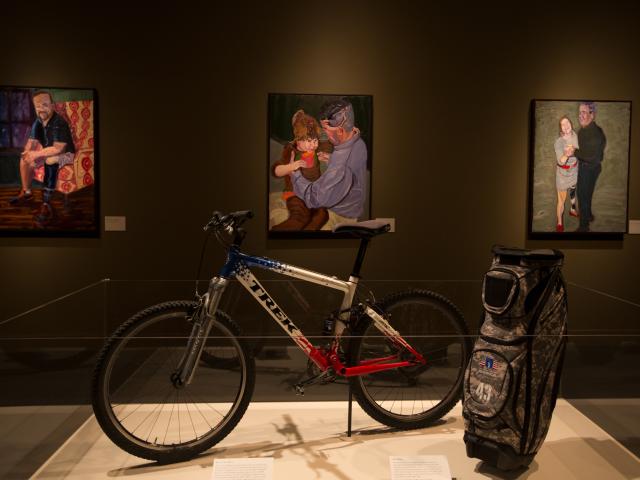 A photo of a bicycle and golf bag display surrounded by portraits painted by President George W Bush, part of the 2017 exhibit Portraits of Courage, at the George W Bush Library and Museum