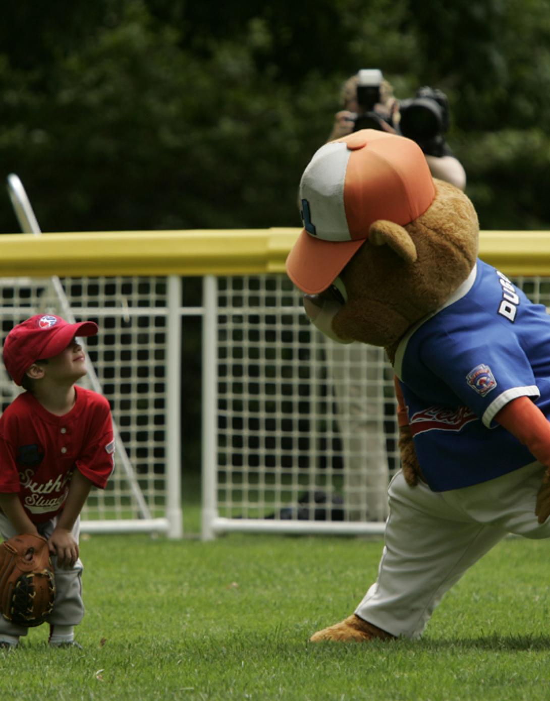 A tee ball player smiles at the mascot in the out field.
