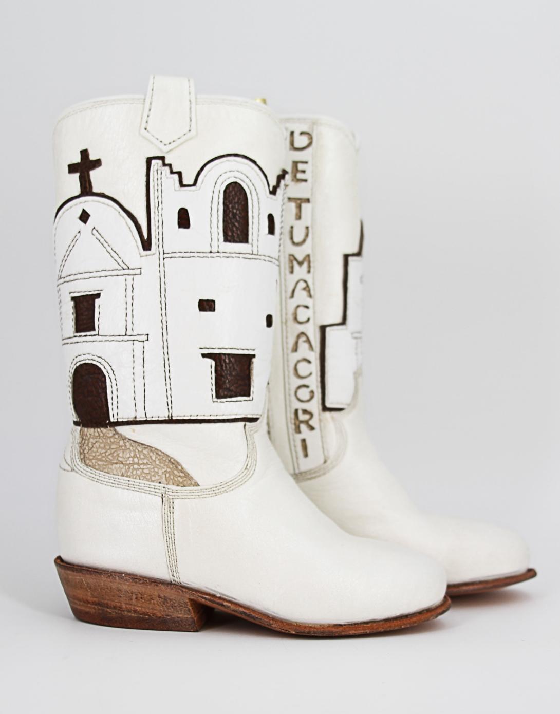 Miniature white leather boots with cutout name, "San Cayentano de Tumacacori," leatherwork depiction of the mission, and a gold leather cord for hanging. Signed inside by master craftsman Paul Bond, AZ, of the Paul Bond Boot Company.